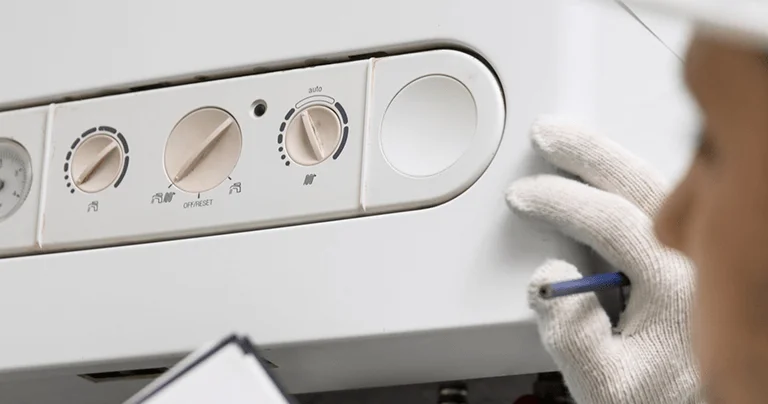 A technician adjusts the control panel of a heating system, featuring knobs and a pressure gauge, while holding a clipboard with a pen.