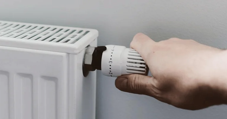 A hand adjusts the thermostat valve on a white radiator, set against a light-colored wall.