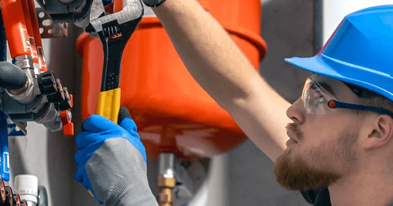 A person in safety gear works on colorful pipes and valves, using an adjustable wrench in a well-lit industrial setting.