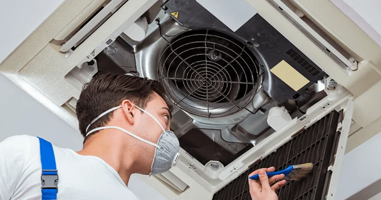 A man in overalls and a mask repairs an air conditioner, focused on his task in a well-lit indoor setting.