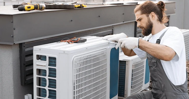 A man repairs an air conditioner, focused on the unit with tools in hand, wearing safety gloves and a work uniform.
