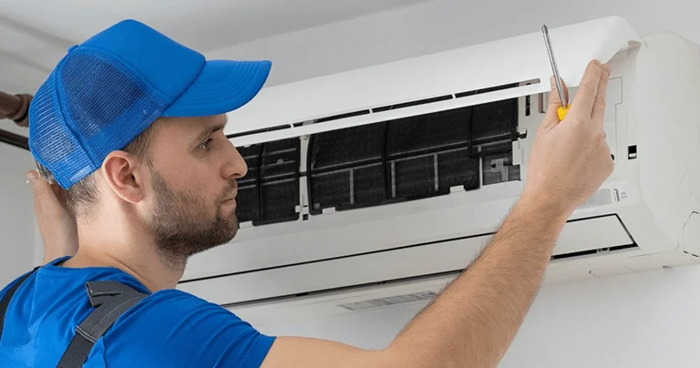 A man in overalls and a cap repairs an air conditioner, focused on his task in a well-lit room.