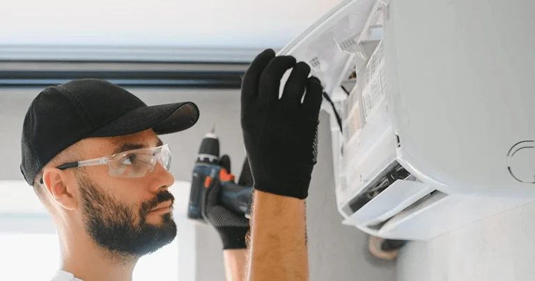 A man wearing overalls and a hat is working on an air conditioner, tools in hand, in a residential setting.