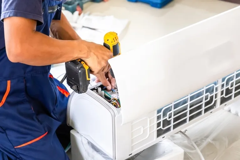 Technician in blue uniform using a yellow cordless drill to repair a white air conditioning unit.