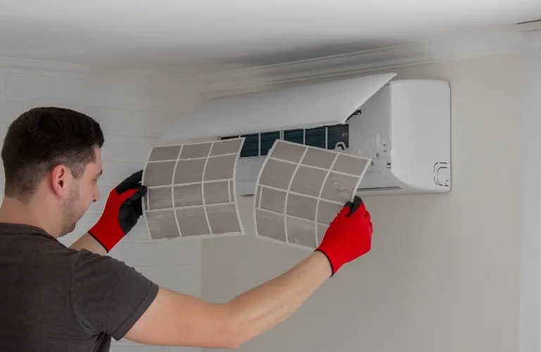 Man wearing red gloves cleaning or replacing filters in a white wall-mounted air conditioning unit.