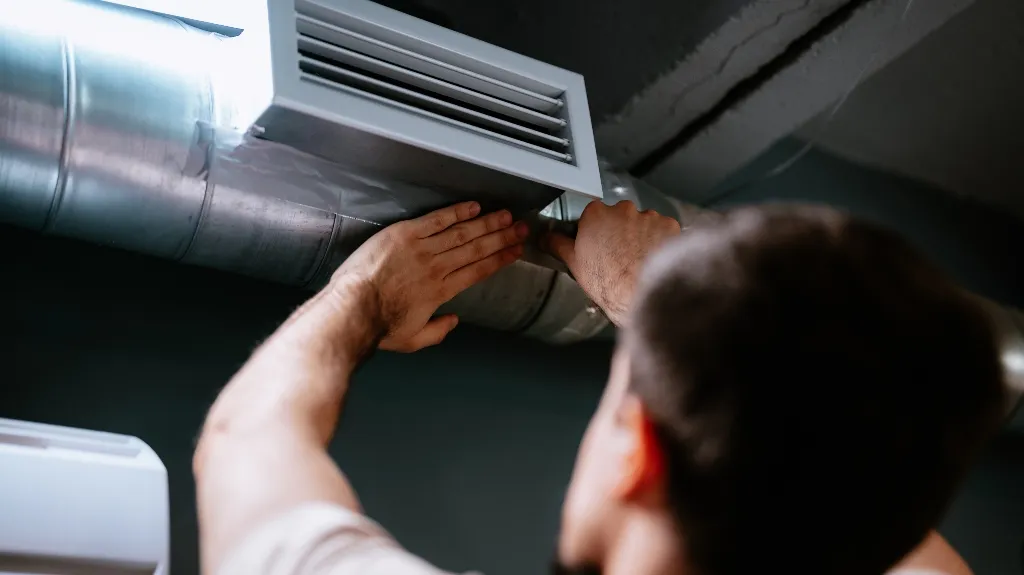 Technician installing or repairing a silver ventilation duct with a white vent cover indoors.