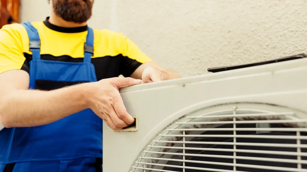 Technician in yellow shirt and blue overalls handling a white air conditioning unit outdoors.