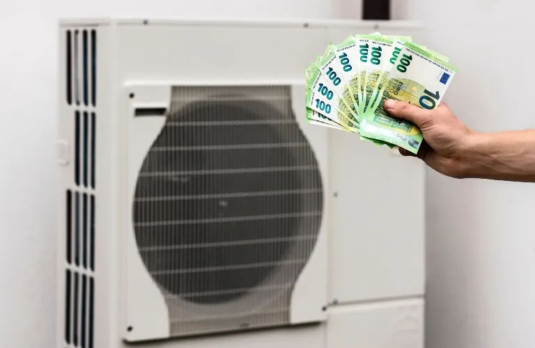 Hand holding multiple 100 and 10 euro banknotes in front of a white air conditioning unit.