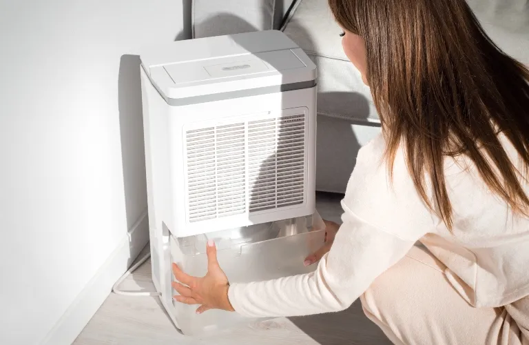 Woman in light clothing emptying water tank from white dehumidifier in bright room.