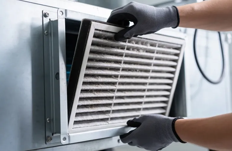 Person wearing gray gloves removing a dusty air filter from a metal HVAC unit.