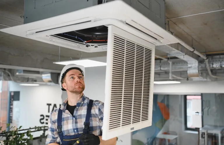 Technician wearing a white hard hat and plaid shirt inspecting a ceiling air conditioning unit.