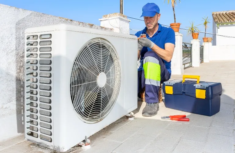 Technician in blue uniform repairing white outdoor air conditioning unit on rooftop with toolbox nearby.