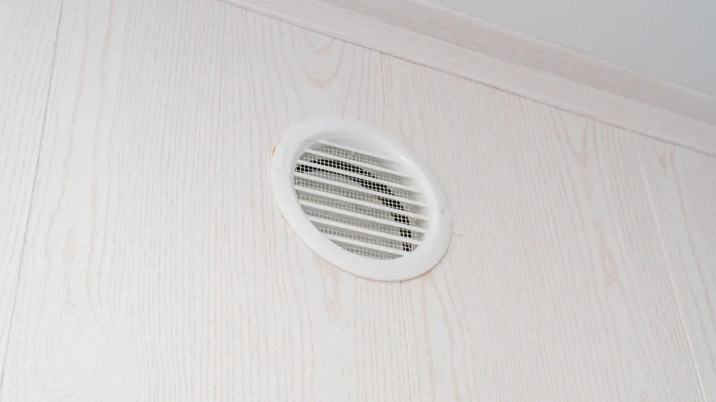 White circular air vent with horizontal slats mounted on a light wood-paneled wall.
