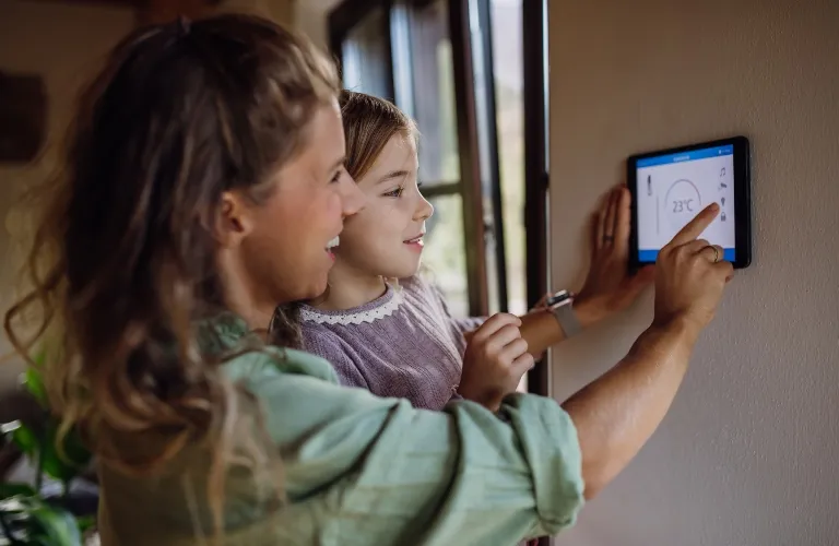 Woman and young girl adjusting a digital thermostat on a beige wall indoors.