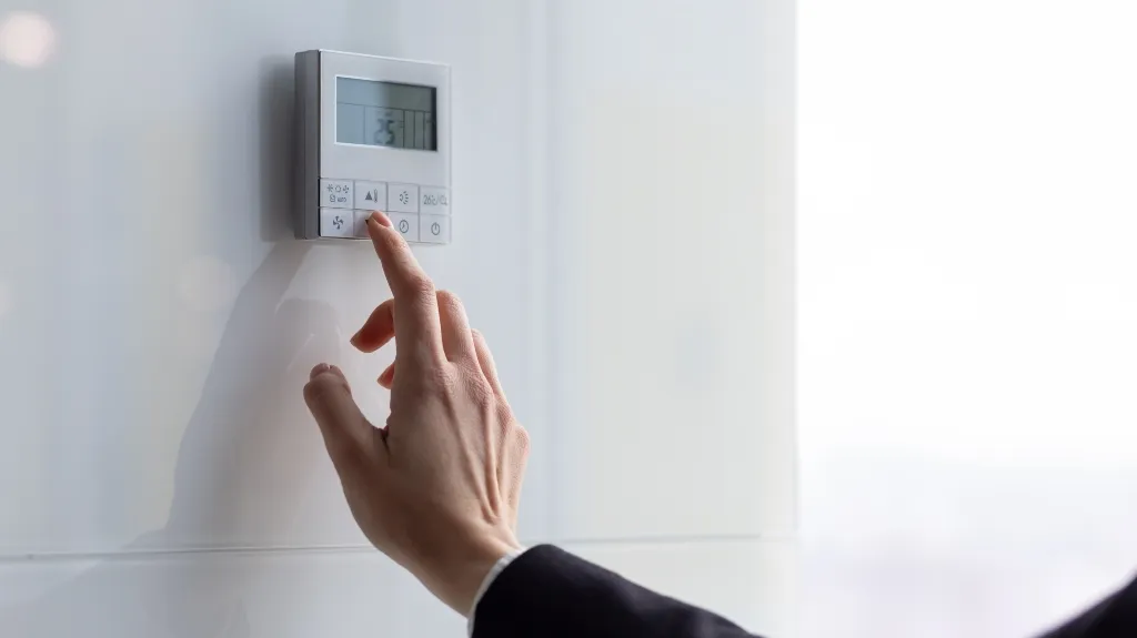 Hand adjusting a white digital thermostat mounted on a light-colored wall.