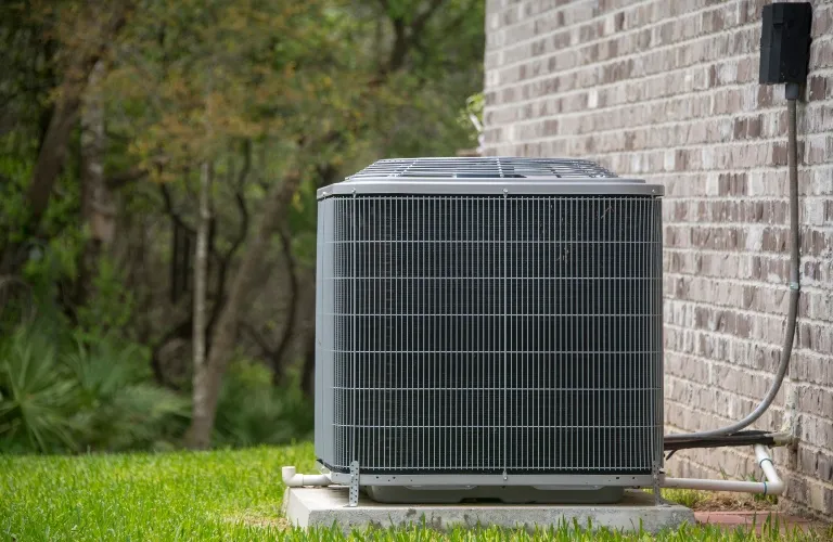 Outdoor gray air conditioning unit installed beside a brick house wall with green grass and trees in the background.