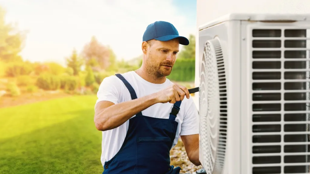Technician in blue cap and overalls repairing outdoor air conditioning unit.