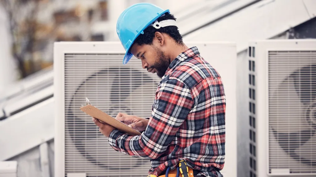 Technician in blue hard hat and plaid shirt inspecting HVAC units with clipboard.